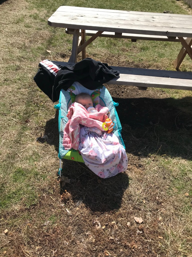 7 1/2 month old baby girl sitting in bouncer covered in blankets and her mamas jacket blocking the sun from her light blonde head while she drinks a bottle near the picnic table in the back yard