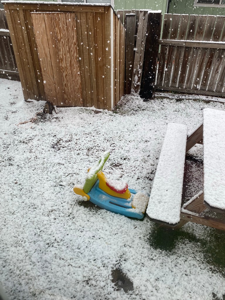 Freshly falling snow on toddler toy, picnic table, and chicken coop in backyard during mid April.