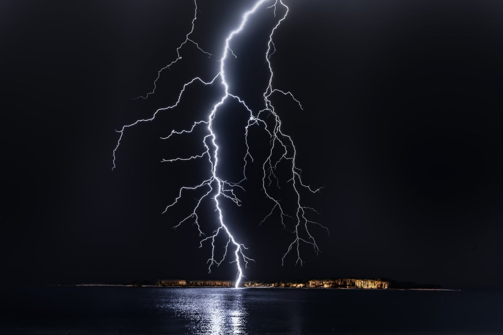 White lightning bolt touching ground near water against a black sky, only the lightning and its reflection on the water are visible 