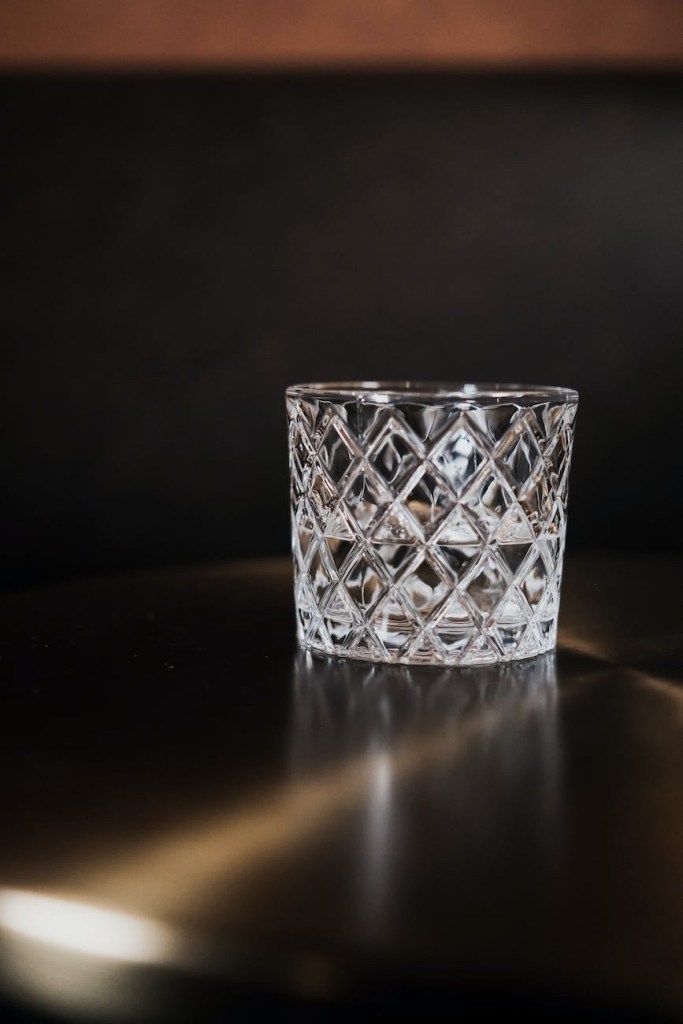 An empty crystal hard alcohol glass against a dark background with a sharp raw of light bouncing off the table top in front of it. 