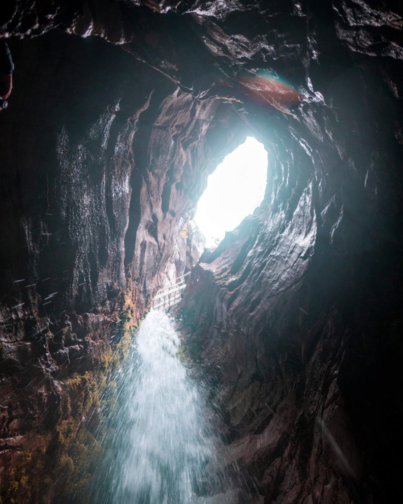 Bright sunlight shining through the exit to a cave with water rushing down.