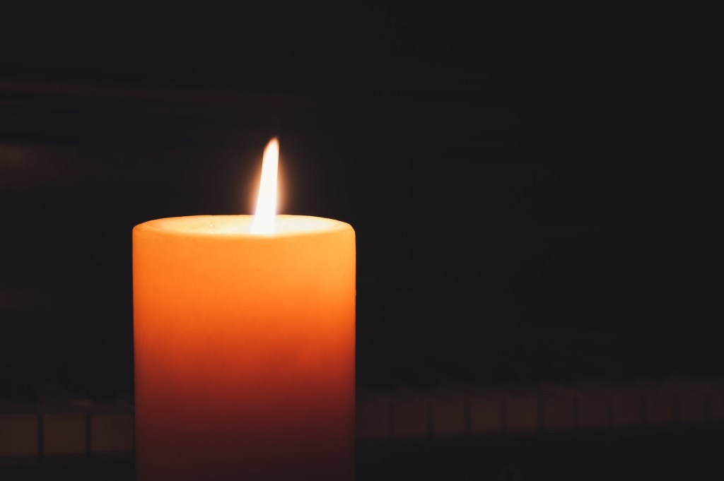 A burning orange wax candle sitting in the foreground of a black background, with faint view of piano keys behind it
